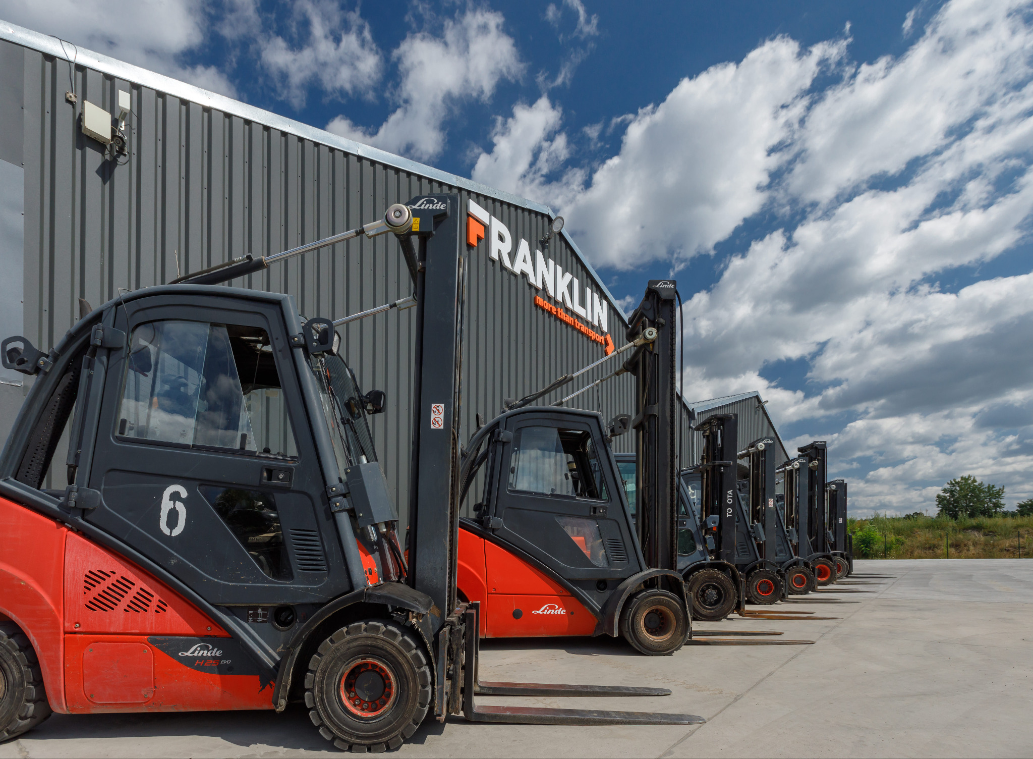 Rząd wózków widłowych Linde przed magazynem z logo Franklin na tle błękitnego nieba z chmurami.