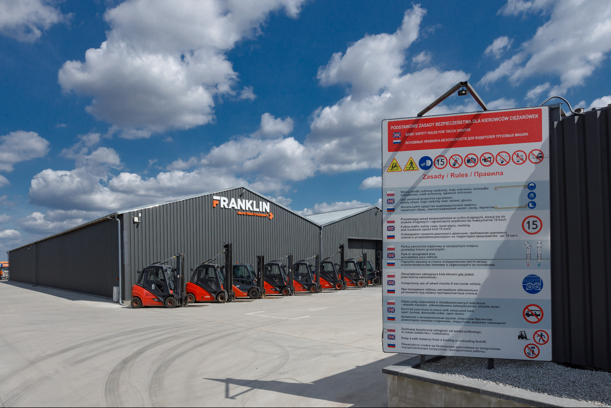 Rząd wózków widłowych Linde zaparkowanych przed szarym magazynem Franklin z tablicą zasad bezpieczeństwa dla kierowców ciężarówek.