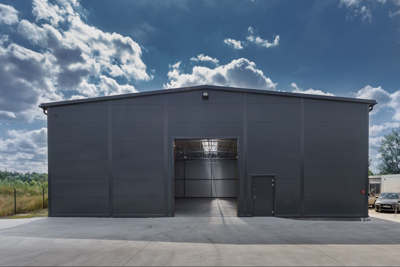 Nowoczesny, szary budynek magazynowy z otwartą bramą wjazdową i małym wejściem bocznym, betonowy plac przed budynkiem, błękitne niebo z chmurami w tle.