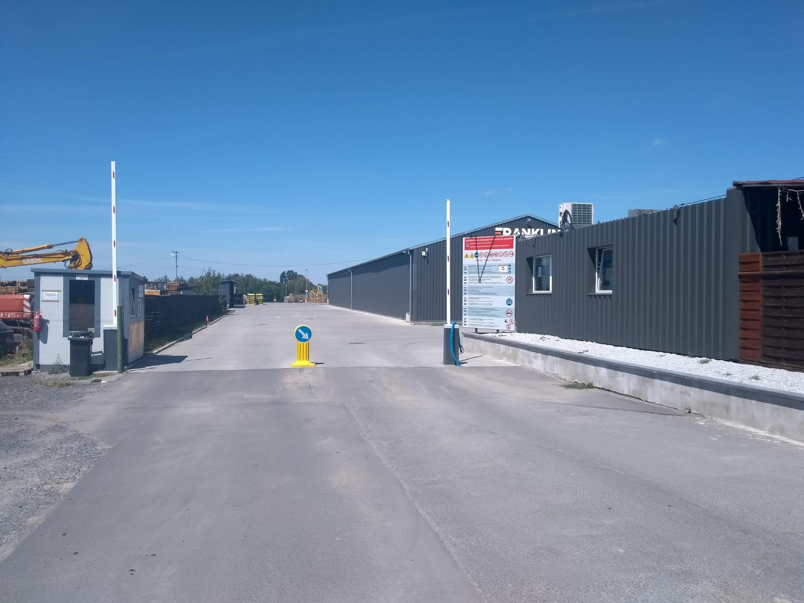 Wjazd na teren magazynowy z budynkami magazynowymi w tle, szlabanem i budką strażniczą po lewej stronie, znak drogowy na środku drogi.