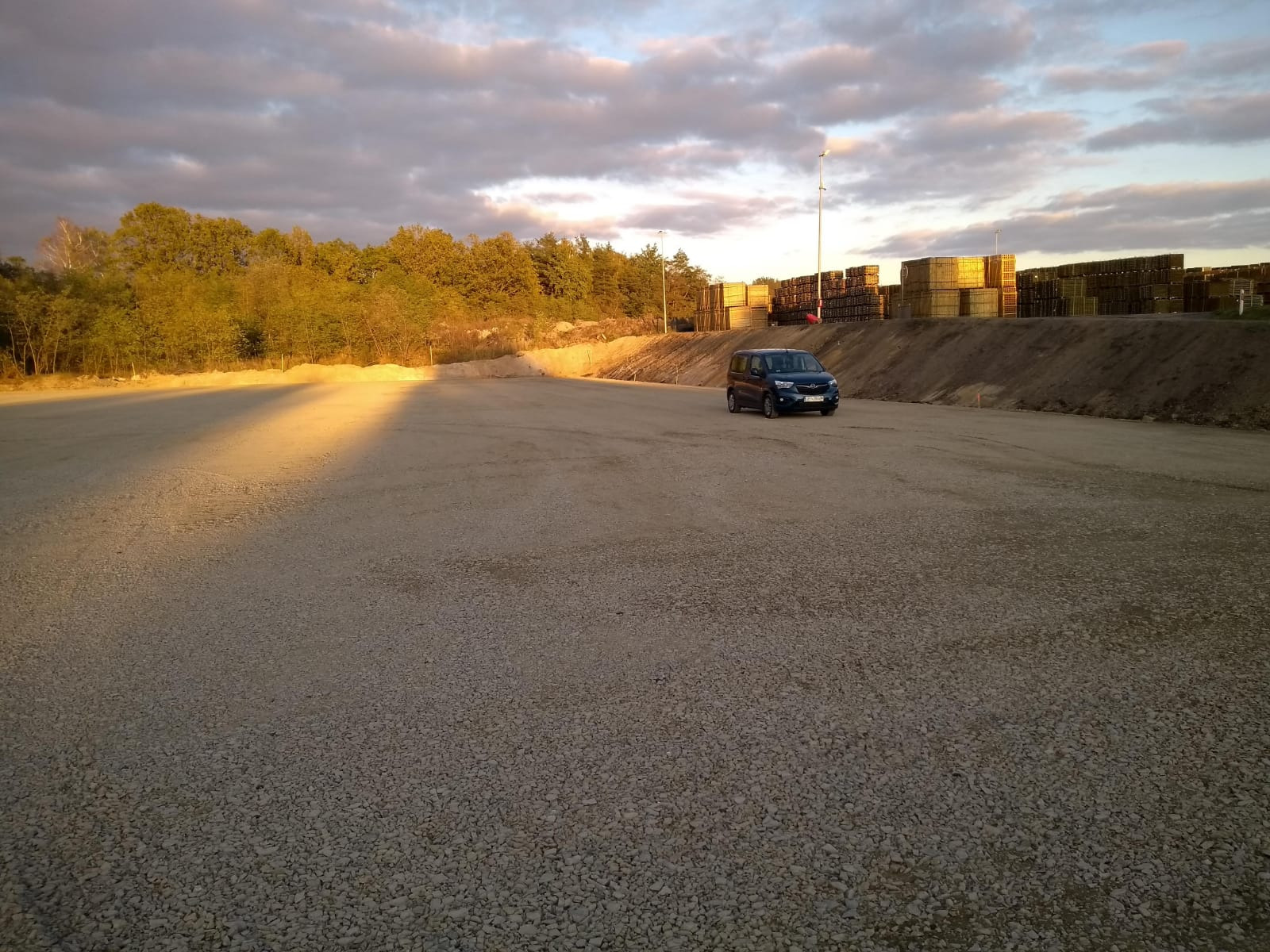 Przygotowany plac pod budowę magazynu z zaparkowanym samochodem dostawczym na tle składowanych palet i zachodzącego słońca.