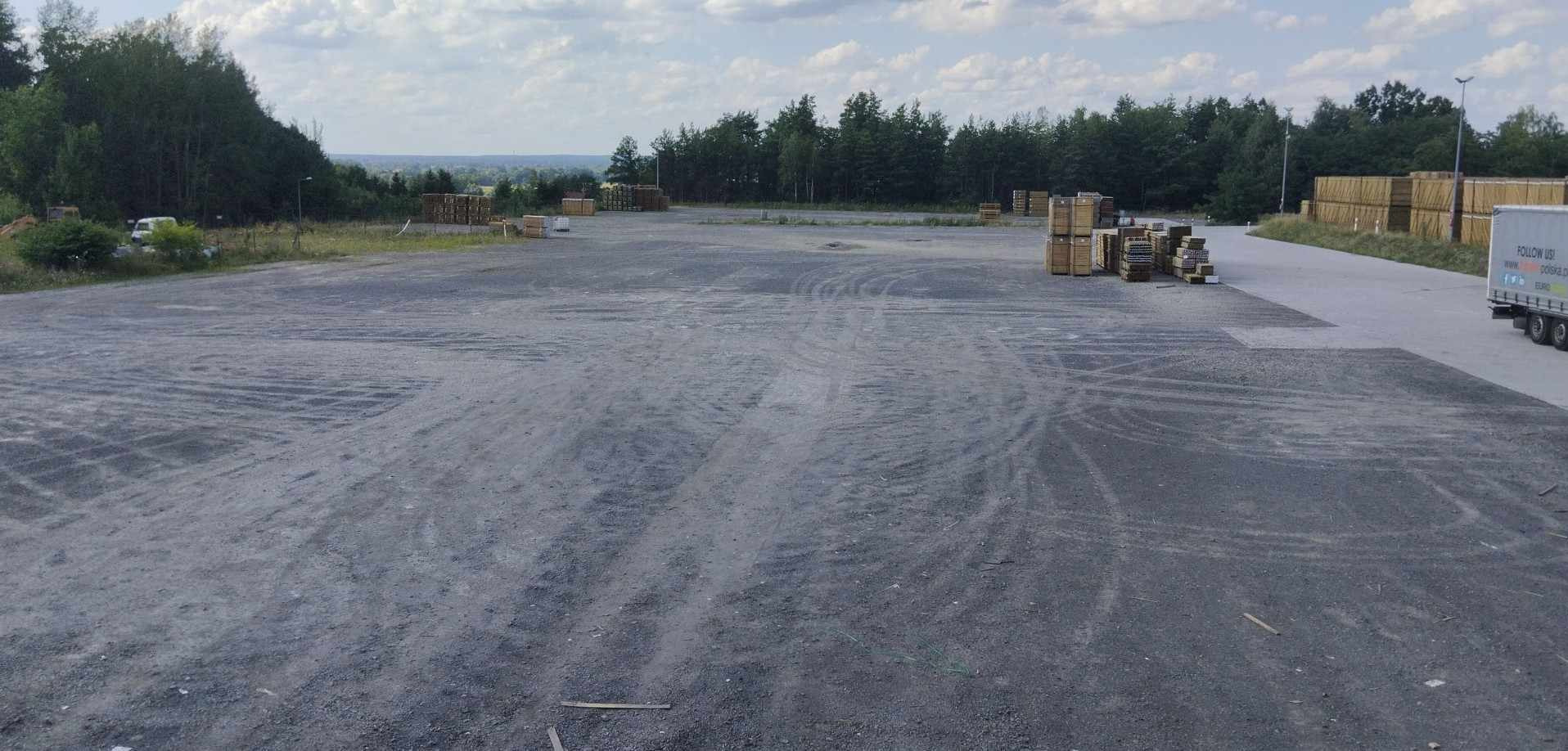 Rozległy plac składowy z ułożonymi paletami i drewnianymi skrzyniami, widoczne ślady opon, w tle zieleń drzew i fragment naczepy ciężarówki.