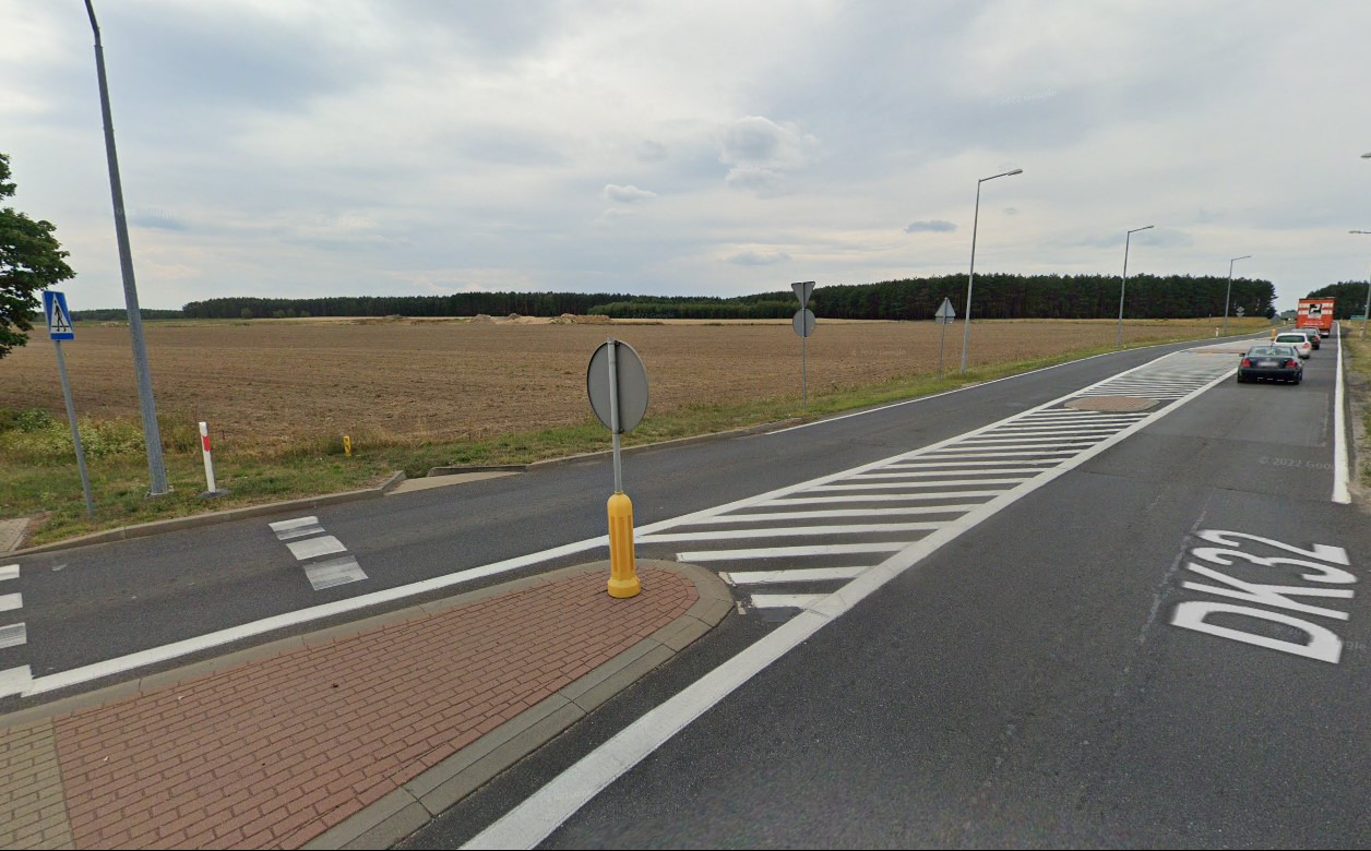 Droga DK32 z pasami dla pieszych w kierunku pola i lasu, widoczne znaki drogowe i słupki, pochmurne niebo.