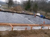 Przygotowanie podłoża pod fundament z widoczną folią izolacyjną, szalunkiem z drewnianych belek i pracownikami w tle. Ujęcie z góry, widoczny krajobraz z drzewami.