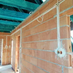 Surowe wnętrze w trakcie prac: pomarańczowe ściany z cegły, niebieskie drewniane belki stropowe, białe przewody poprowadzone po ścianach, puszki instalacyjne zamontowane w ścianie.