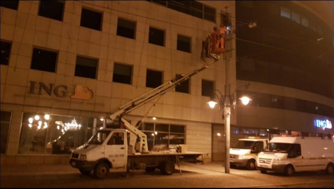 Pracownicy na podnośniku koszowym przy budynku ING, wykonujący prace elektryczne przy elewacji, z widocznymi przewodami i lampami, w tle zaparkowane samochody dostawcze.