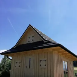 Niewielki, surowy drewniany domek z czarnym dachem, stylizowanymi okiennicami i przeszklonymi drzwiami wejściowymi, na tle błękitnego nieba i zielonego krajobrazu.