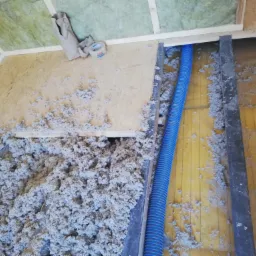 Poddasze w trakcie izolacji: luźna wełna mineralna wypełnia przestrzeń między krokwiami, widoczny fragment niebieskiego przewodu wentylacyjnego i rolka taśmy klejącej na płycie OSB.