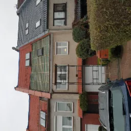 Dom w zabudowie szeregowej z częściowo zdjętym czerwonym dachem, widoczne przygotowanie pod nowe pokrycie, niebieski samochód zaparkowany przed budynkiem.