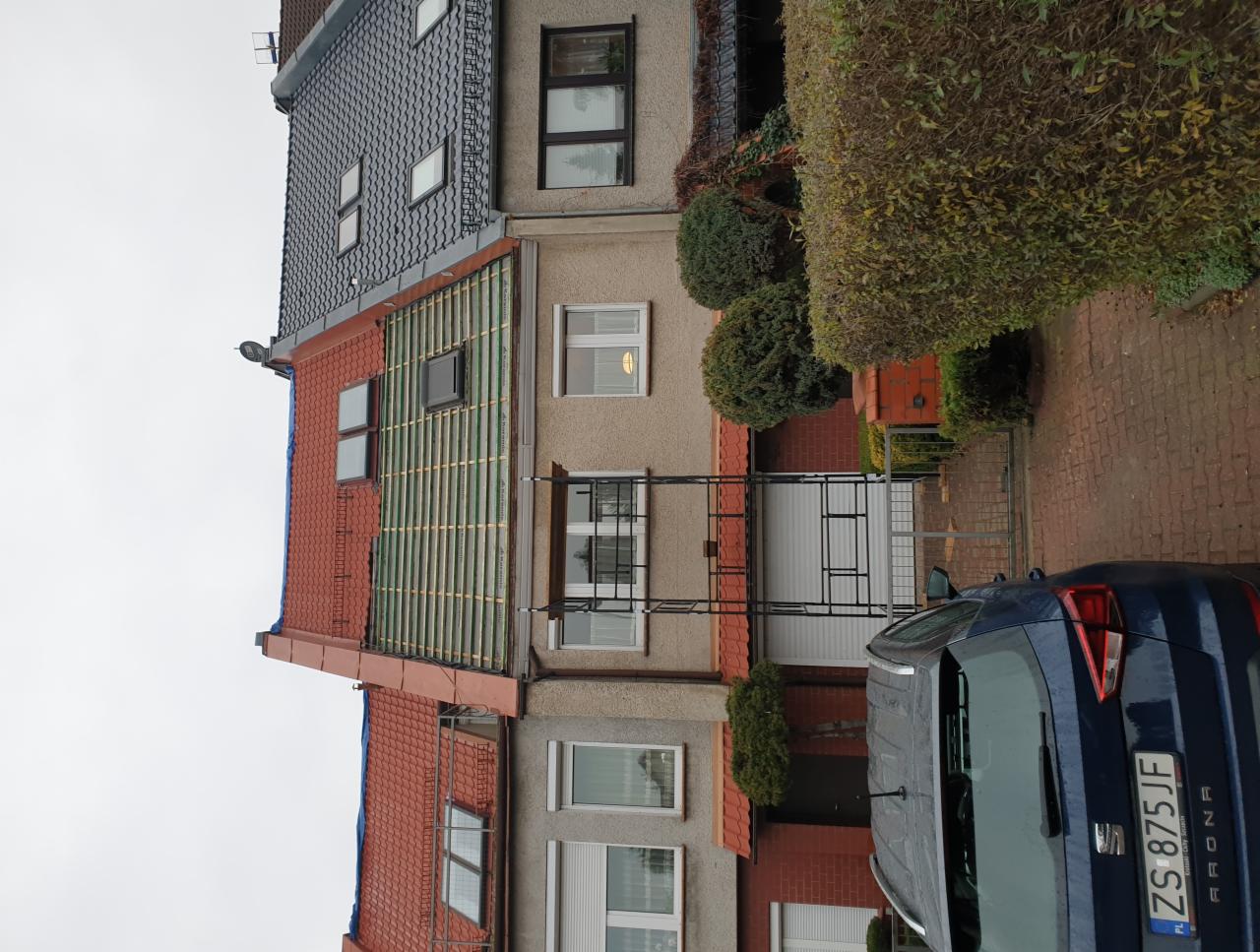 Dom w zabudowie szeregowej z częściowo zdjętym czerwonym dachem, widoczne przygotowanie pod nowe pokrycie, niebieski samochód zaparkowany przed budynkiem.