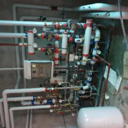 Złożona instalacja grzewcza z licznymi rurami, zaworami i manometrami, zrealizowana w Białystoku, widoczna z perspektywy sufitu.