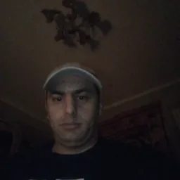 Portret mężczyzny w czarnej koszulce i beżowej czapce z daszkiem, z lampą w tle, w słabym oświetleniu.