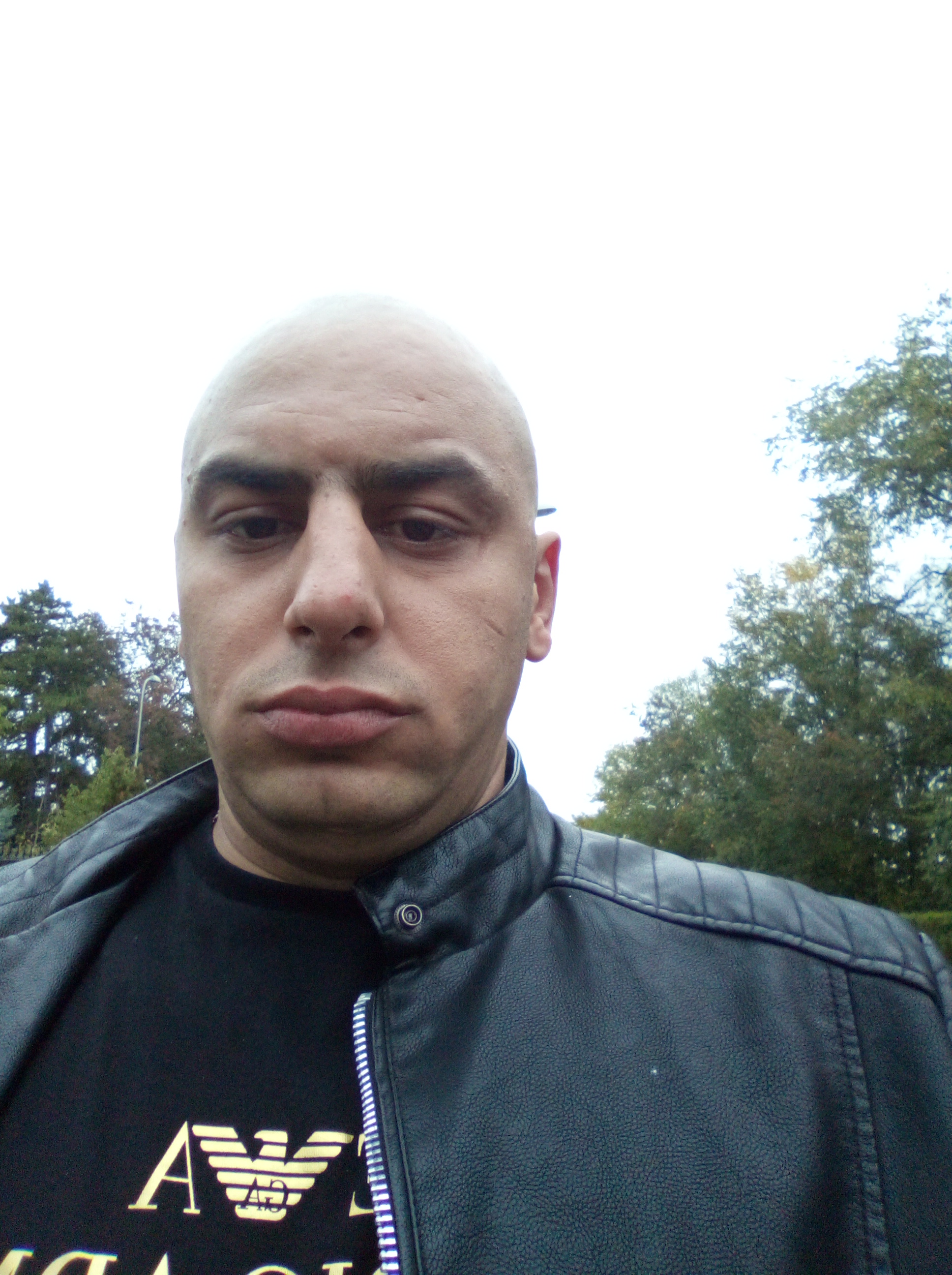 Mężczyzna z ogoloną głową w czarnej skórzanej kurtce i koszulce z logo na tle drzew i jasnego nieba. Zdjęcie z bliska.