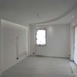 Wnętrze pokoju w trakcie remontu: ściany po gładziowaniu, zabezpieczone okno, widoczne przewody elektryczne na suficie, jasne panele podłogowe.