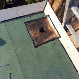 ESPANIOL-BUD - Fragment dachu płaskiego z zieloną papą, drewnianą obróbką krawędzi i odpływem zabezpieczonym masą bitumiczną; widoczne ślady po wierceniu w papie i kilka wkrętów na powierzchni.