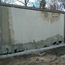 ESPANIOL-BUD - Elewacja budynku w trakcie naprawy, z widocznymi fragmentami starego tynku, nałożonym gruntem i przygotowanym podłożem, z rynną w górnej części i ziemią u podstawy ściany.