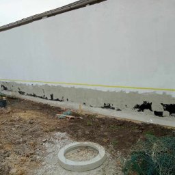 ESPANIOL-BUD - Przygotowanie elewacji budynku: gruntowanie ściany z widocznymi ubytkami, żółta taśma wyznaczająca linię, ziemia i materiały budowlane w tle.