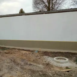 Świeżo pomalowana biała elewacja budynku z widocznym gruntowaniem dolnej części, zabezpieczona żółtą taśmą malarską, na pierwszym planie rozkopana ziemia i betonowy krąg.