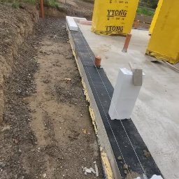 ESPANIOL-BUD - Fundament budynku w trakcie budowy: widoczne bloczki Ytong, sznurek murarski, poziomica, gumowy młotek na ziemi, ocieplenie fundamentu, w tle dom jednorodzinny.