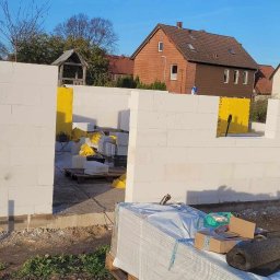 ESPANIOL-BUD - Wznoszenie ścian domu z bloczków betonowych, widoczne materiały budowlane, izolacja w żółtych workach, w tle zabudowa jednorodzinna.