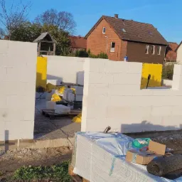 Wznoszenie ścian domu z bloczków betonowych, widoczne materiały budowlane, izolacja w żółtych workach, w tle zabudowa jednorodzinna.