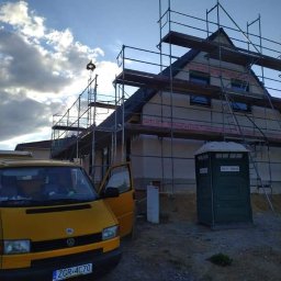 ESPANIOL-BUD - Dom jednorodzinny w trakcie prac elewacyjnych, otoczony rusztowaniem, z widoczną żółtą furgonetką i przenośną toaletą na placu budowy, przy zachmurzonym niebie.