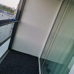 Wnętrze balkonu z szarą wykładziną dywanową, białą ścianą i oknem z widokiem na miasto w oddali.