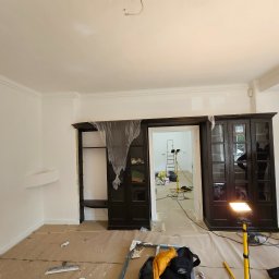 ESPANIOL-BUD - Malowanie mieszkania: pokój w trakcie odświeżania, ściany pomalowane na biało, podłoga zabezpieczona papierem, widoczne narzędzia malarskie i oświetlenie halogenowe, fragment szafy zabezpieczony...