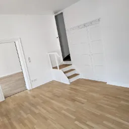 Jasne panele podłogowe w pustym, odnowionym pokoju z białymi ścianami, schodami i drzwiami.