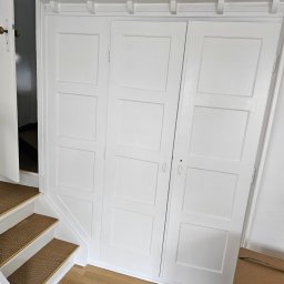 ESPANIOL-BUD - Biała, zabudowana szafa wnękowa z przesuwnymi drzwiami i ozdobnym gzymsem, zlokalizowana przy schodach z wykładziną w kolorze beżowym. Widoczna otwarta boczna szafka oraz karton na podłodze.