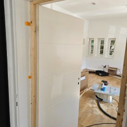 ESPANIOL-BUD - Montaż drzwi wewnętrznych: białe, nowoczesne drzwi z poziomymi frezowaniami w surowej drewnianej ościeżnicy, widoczne narzędzia i resztki drewna na podłodze w jasnym pomieszczeniu z trzema oknami.