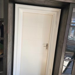 ESPANIOL-BUD - Montaż białych drzwi wewnętrznych z poziomymi panelami, widoczna uszczelka silikonowa, śrubokręt oraz drewniana rama w ciemnym kolorze.