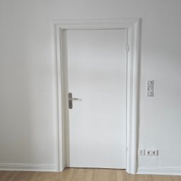 ESPANIOL-BUD - Białe, wewnętrzne drzwi z poziomymi wgłębieniami, zamontowane w białej ościeżnicy, na tle białej ściany z gniazdkami i włącznikami, oraz podłogi z paneli w odcieniach drewna.