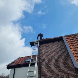 ESPANIOL-BUD - Pracownik na drabinie naprawia dach z czerwonej dachówki, widoczne ceglana ściana budynku i błękitne niebo z chmurami.