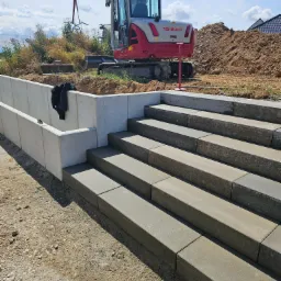 Budowa schodów zewnętrznych z betonowych elementów modułowych, widoczny minikoparka Takeuchi i wykop ziemi w tle.