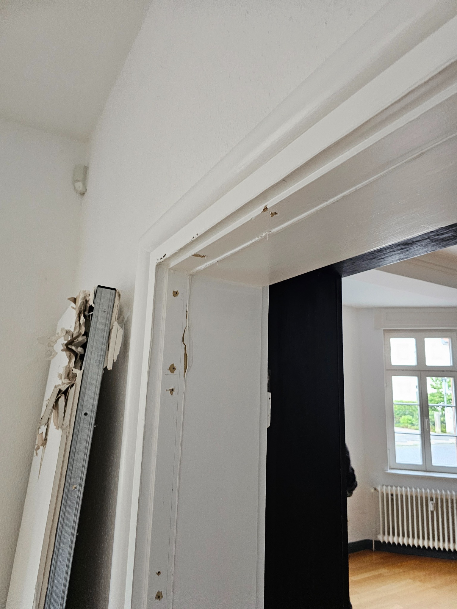 Demontaż ościeżnicy drzwiowej, widoczne uszkodzenia i resztki farby na ścianie oraz elementy starej ościeżnicy oparte o ścianę.