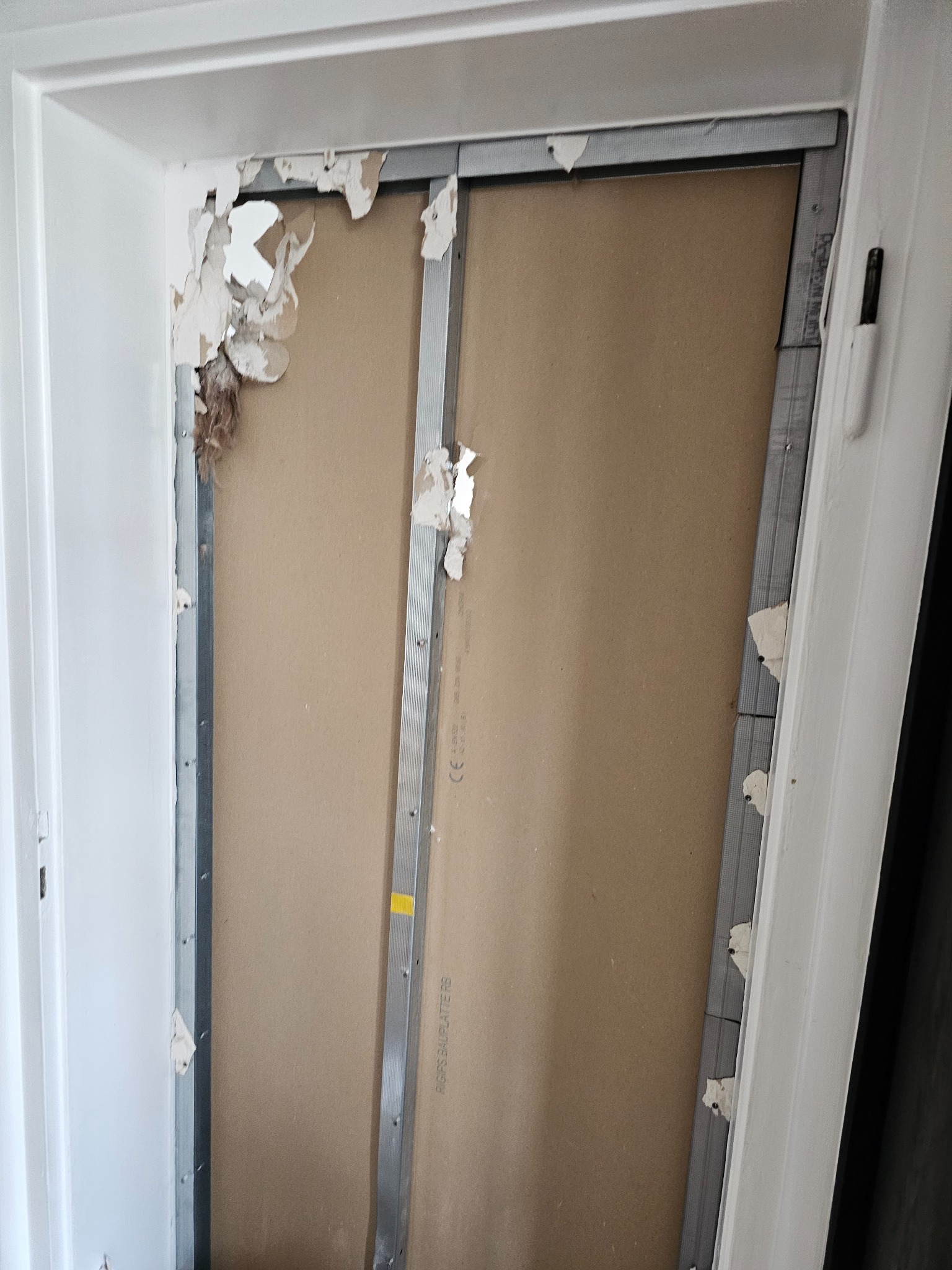 Fragment ściany z uszkodzonym tynkiem, odsłoniętą konstrukcją metalową i płytą gipsowo-kartonową w trakcie prac remontowych, widoczne resztki starego wykończenia i przygotowanie do dalszych etapów.