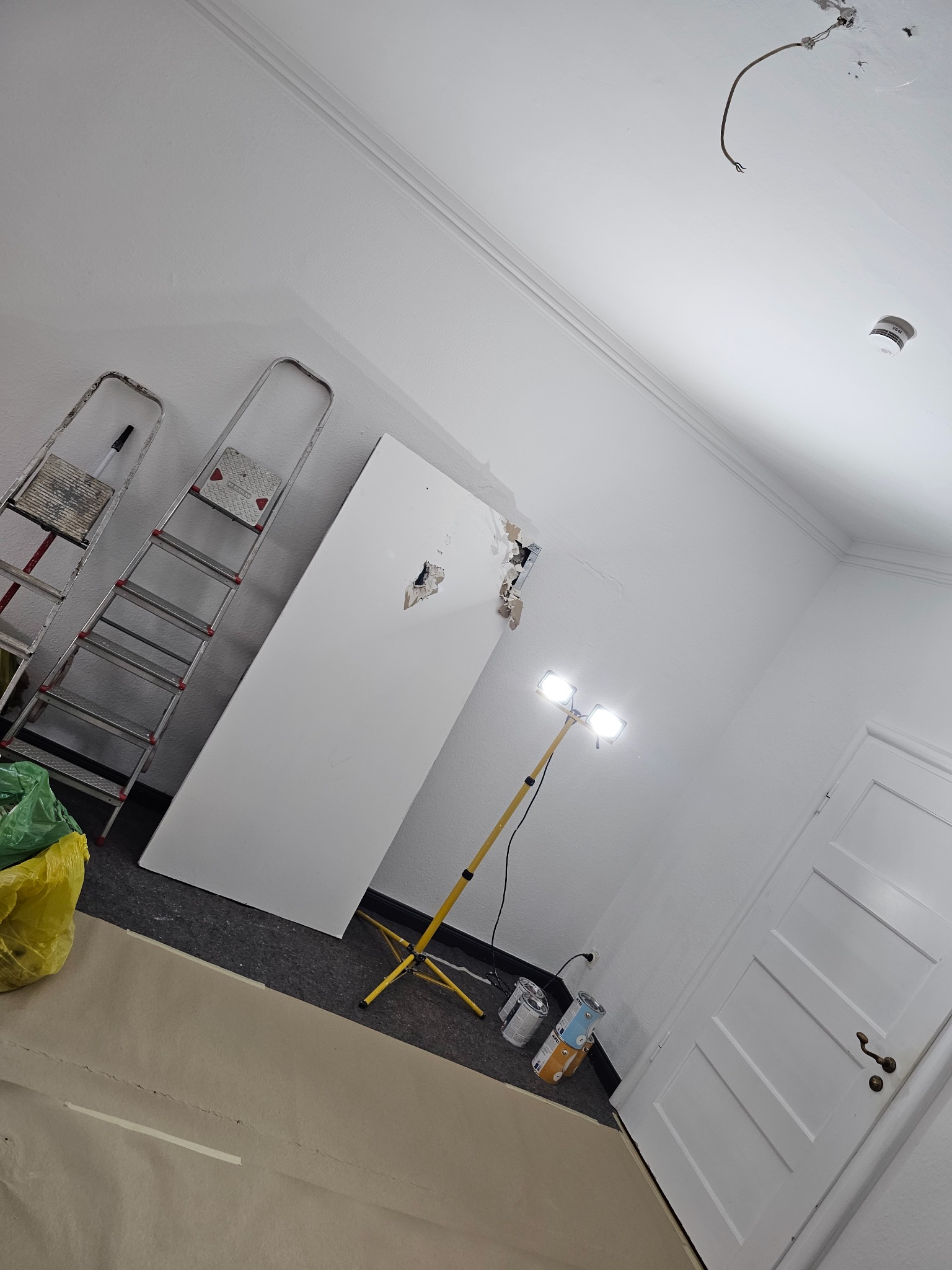 Wnętrze w trakcie remontu: ściana z uszkodzonym tynkiem, drabiny oparta o ścianę, reflektor halogenowy na statywie oświetla uszkodzone miejsce, podłoga zabezpieczona papierem malarskim, widoczne...
