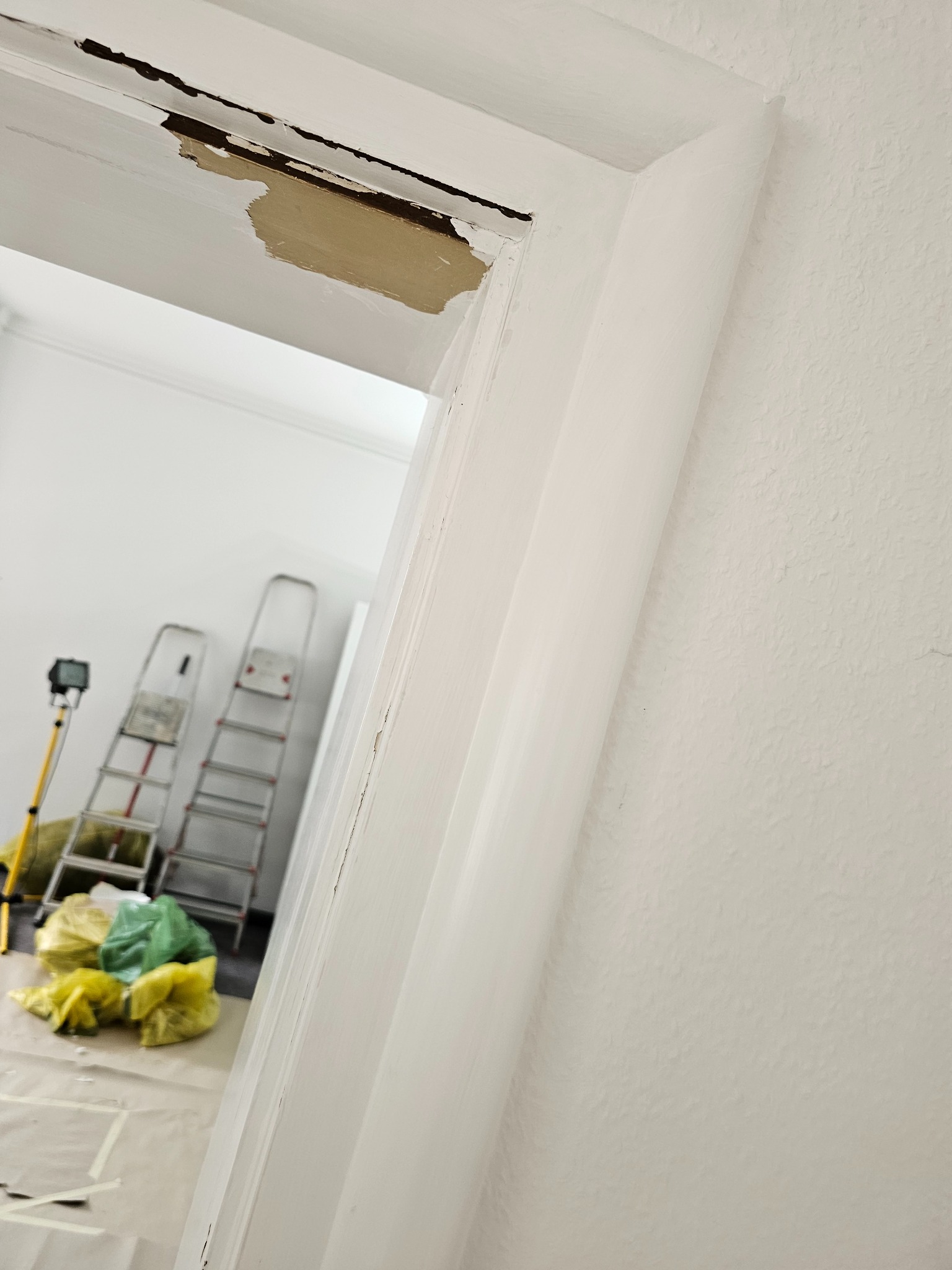 Fragment ościeżnicy drzwiowej w trakcie renowacji, z widocznymi warstwami starej farby i przygotowaniem do malowania; w tle drabiny i worki z odpadami.