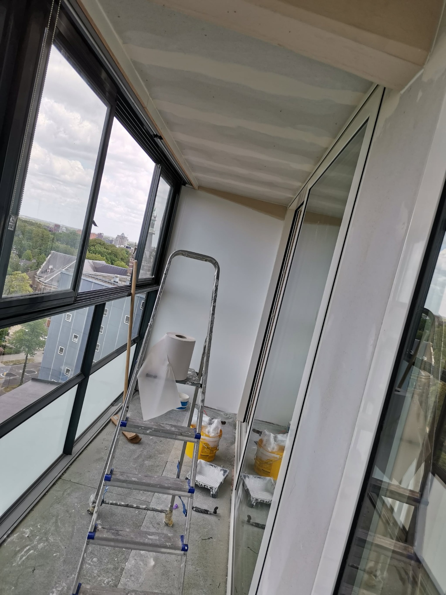 Balkon w trakcie remontu: aluminiowa drabina z papierem ochronnym, wiaderka z farbą, widok na miasto przez duże okna.