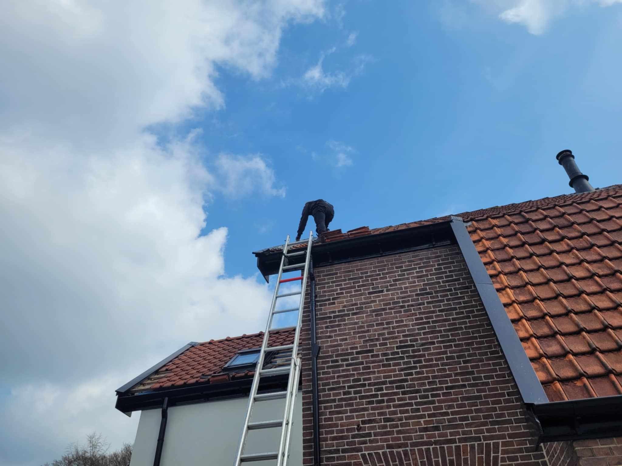 Pracownik na drabinie naprawia dach z czerwonej dachówki, widoczne ceglana ściana budynku i błękitne niebo z chmurami.