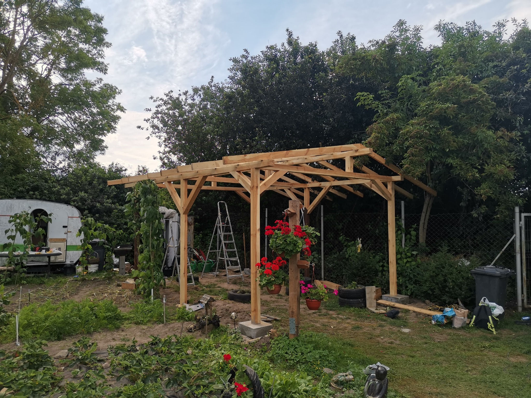 Drewniana konstrukcja altany w trakcie budowy, widoczna drabina oparta o jeden ze słupów, w tle przyczepa kempingowa i bujna zieleń ogrodu.