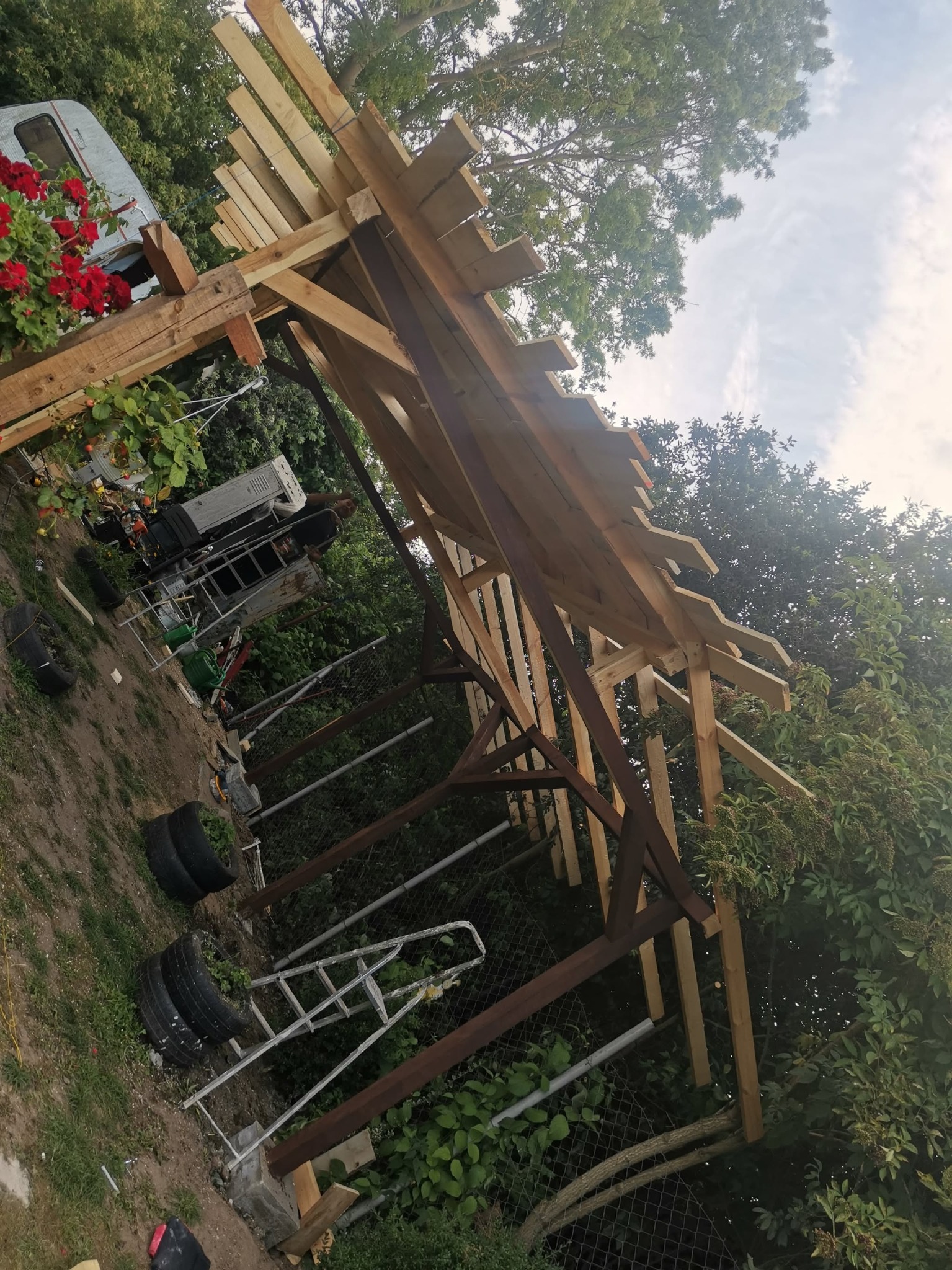 Konstrukcja drewniana altany w trakcie budowy w Baniach, widoczne elementy szkieletu dachu i słupy nośne, w tle przyczepa kempingowa i zieleń.