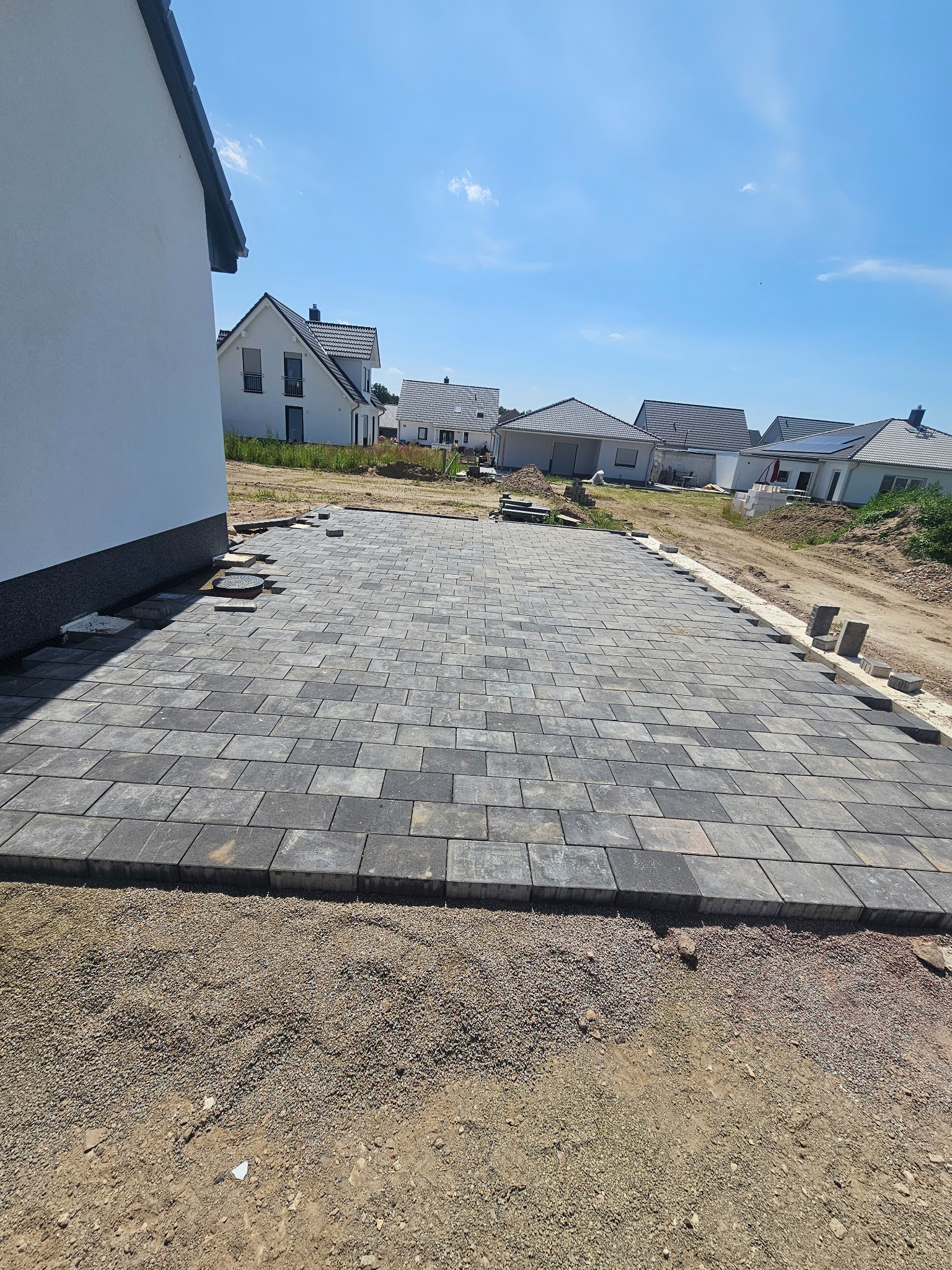 Ułożona kostka brukowa na podwórku nowego domu jednorodzinnego, widoczne przygotowane obrzeża i podsypka, w tle inne domy.