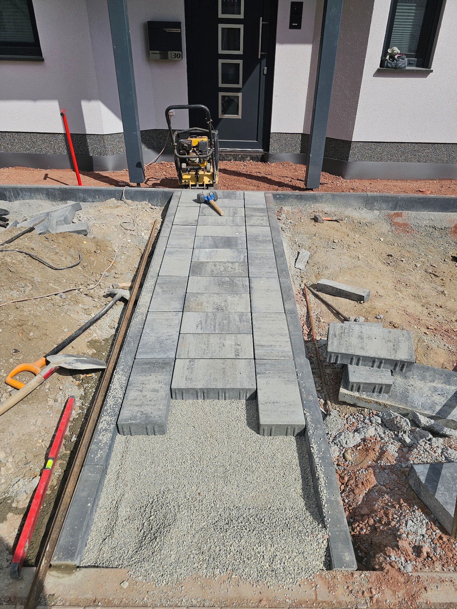 Układanie szarej kostki brukowej na podsypce z kruszywa, widoczny ubijak wibracyjny, poziomica, łopata i fragment elewacji budynku z drzwiami wejściowymi