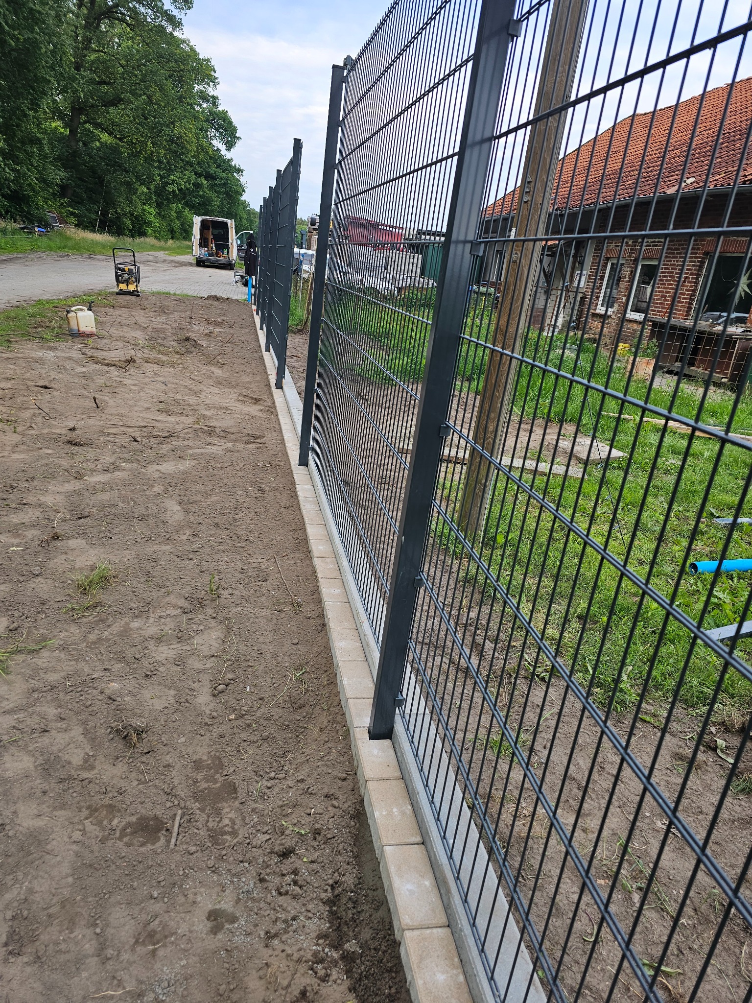 Montaż segmentowego ogrodzenia panelowego na podmurówce z betonowych bloczków, widoczny teren budowy z ubijarką i samochodem dostawczym w tle.