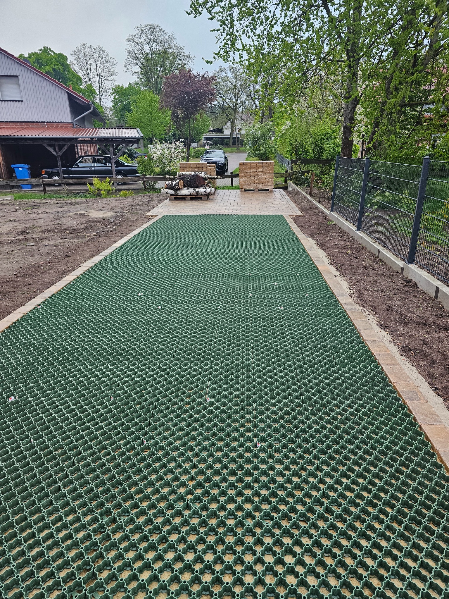 Zielona, ażurowa nawierzchnia trawnikowa z obrzeżami z kostki, częściowo ułożona na przygotowanym podłożu, widok z góry w kierunku domu i podwórza.