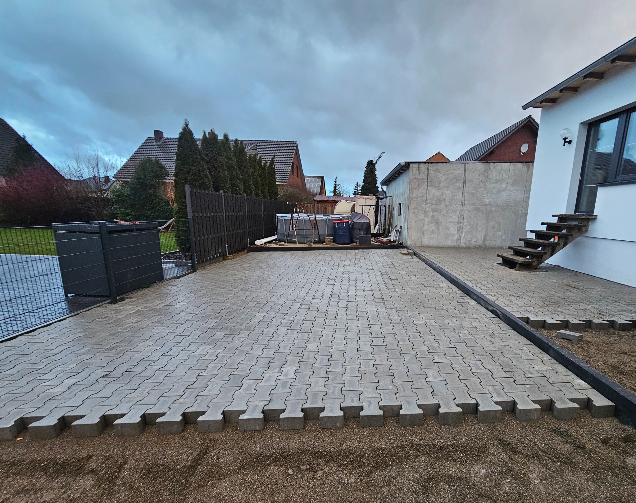 Układanie szarej kostki brukowej na podwórku domu jednorodzinnego, widoczny częściowo ułożony fragment z pozostawionym piaskiem, w tle ogrodzenie, trawnik, basen i budynek gospodarczy, pochmurne...