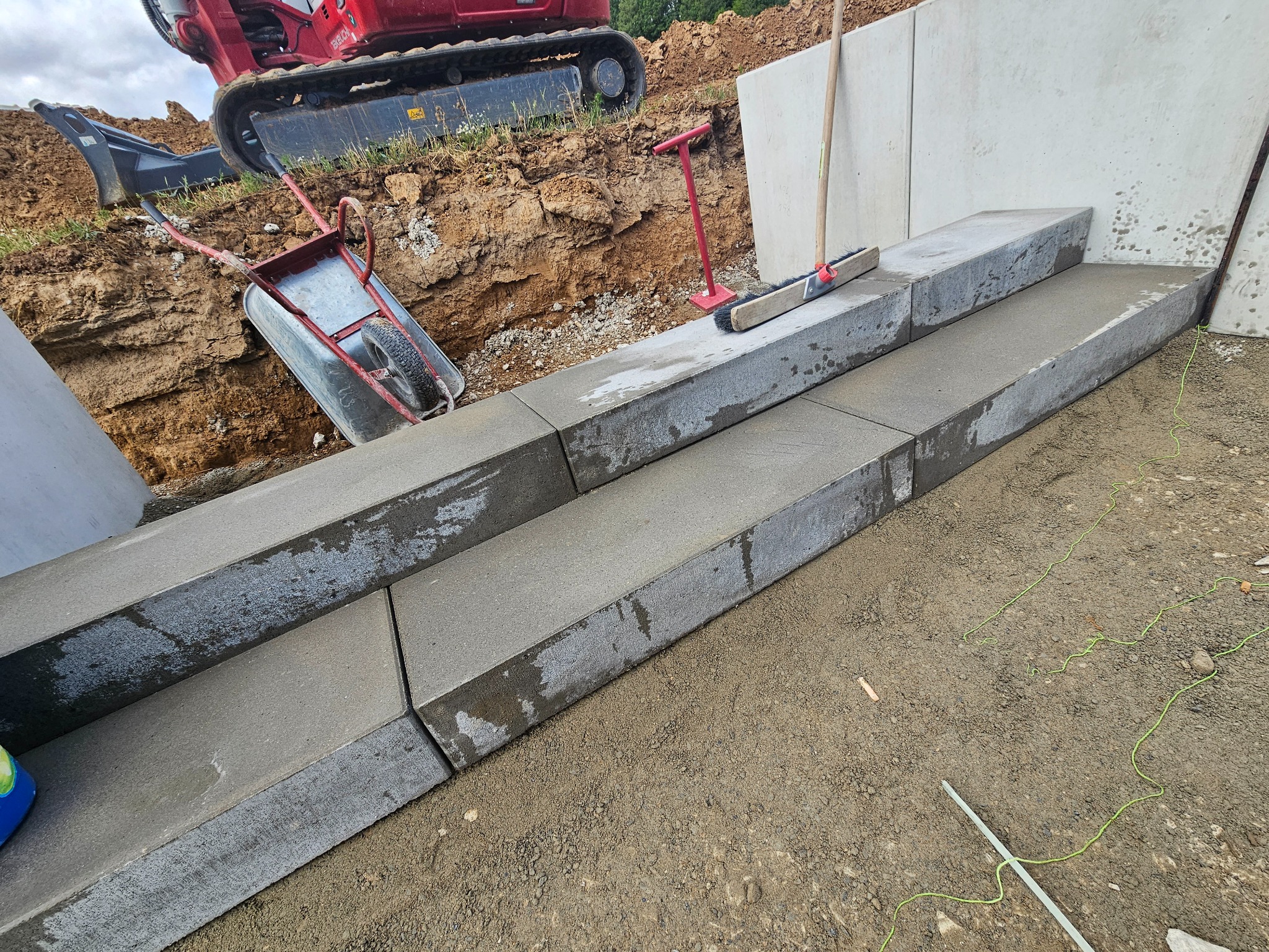 Betonowe schody zewnętrzne w trakcie budowy, widoczny minikoparka, taczka, szczotka i narzędzia na placu budowy.