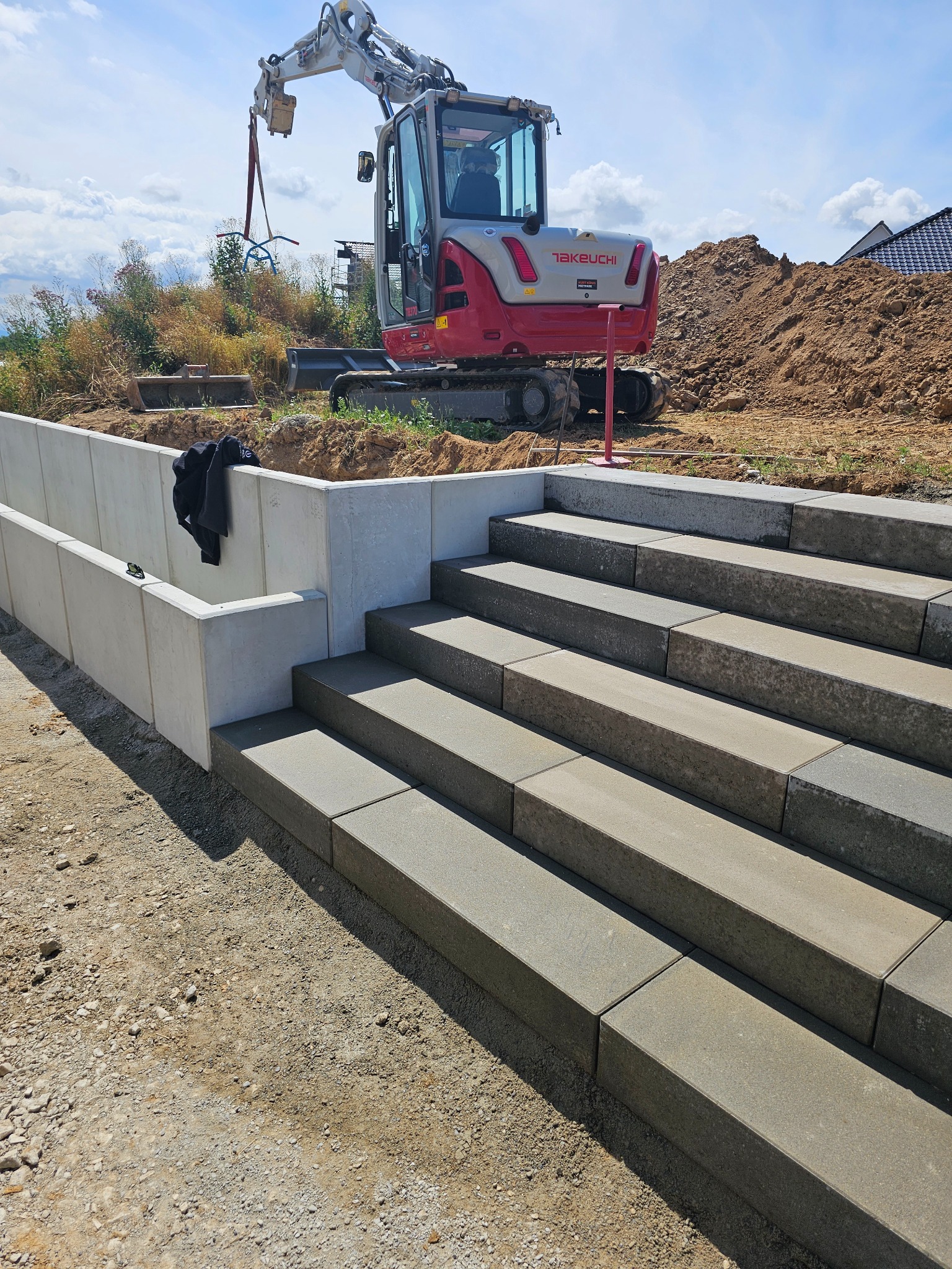 Budowa schodów zewnętrznych z betonowych elementów modułowych, widoczny minikoparka Takeuchi i wykop ziemi w tle.
